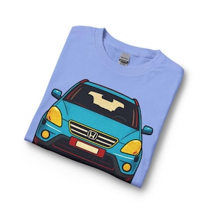 May include: Light blue t-shirt featuring a cartoon illustration of a teal car with yellow headlights, a red license plate, and a black grill. The car graphic is centered on the shirt.