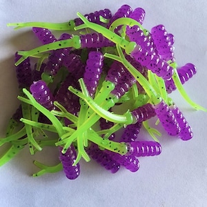 Może przedstawiać: Stos miękkich, plastikowych przynęt wędkarskich. Przynęty są fioletowe z czarnymi plamkami i mają jasnozielone, segmentowane ogony. Przynęty mają naśladować wygląd małego stworzenia wodnego.