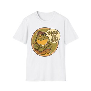 May include: White t-shirt with a cartoon frog wearing a brown hat and vest, arms crossed. The frog is inside a circular design with the text "TOAD YA SO!" in a speech bubble. The design has a vintage, retro style.