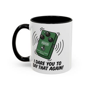 Puede incluir: Taza de cerámica blanca con interior y asa negros. La taza presenta una ilustración verde de un pedal MXR Carbon Copy Delay con el texto "I DARE YOU TO SAY THAT AGAIN!" impreso debajo.