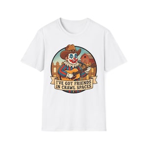 Puede incluir: Camiseta blanca con un payaso de dibujos animados con sombrero de vaquero tocando la guitarra. El payaso está dentro de un diseño circular con un granero y una valla. El texto "I'VE GOT FRIENDS IN CRAWL SPACES" está en una pancarta.