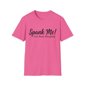 May include: A bright pink t-shirt with the text "Spank Me! I've Been Naughty" in a black script font. The shirt has a crew neck and short sleeves, and is made of a soft material.