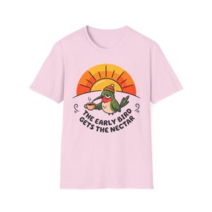 May include: Light pink t-shirt featuring a cartoon hummingbird wearing a knit cap and holding a coffee cup. The bird is set against a rising sun graphic. Text reads "THE EARLY BIRD GETS THE NECTAR."