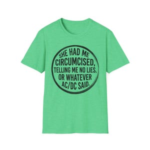 May include: A light green t-shirt with a black circular graphic. The graphic reads "SHE HAD ME CIRCUMCISED, TELLING ME NO LIES. OR WHATEVER AC/DC SAID." The shirt is made of a soft material.