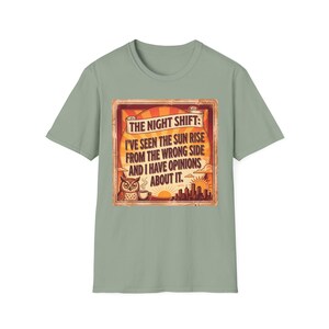 Puede incluir: Camiseta verde salvia con un diseño rectangular. El diseño incluye el texto "THE NIGHT SHIFT: I'VE SEEN THE SUN RISE FROM THE WRONG SIDE AND I HAVE OPINIONS ABOUT IT." Un búho, una taza de café y un paisaje urbano también están presentes.