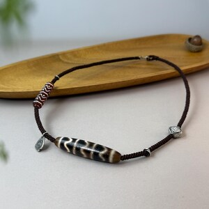 Könnte beinhalten: Eine braune Kordelkette mit einer zentralen zylindrischen Perle mit einem schwarz-weißen Muster. Die Halskette enthält kleinere gemusterte Perlen und einen silberfarbenen Anhänger. Silberfarbene quadratische Akzente sind in das Design integriert.