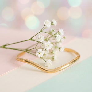 Op de afbeelding: Een goudkleurige, vierkante armband met een golvend ontwerp, rustend op een pastelkleurig oppervlak. Delicate witte bloemen met kleine bloesems zijn bij de armband gerangschikt, wat een zachte, elegante esthetiek creëert.