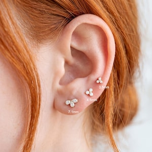 Può includere: Tre orecchini a lobo in oro, ciascuno composto da un gruppo di tre piccole pietre trasparenti. Gli orecchini sono etichettati con le misure 3,5 mm, 4,5 mm e 7,1 mm, che indicano le rispettive larghezze sul lobo e sulla cartilagine.