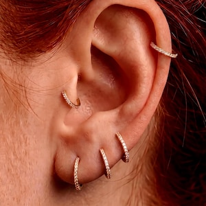 Pode incluir: Close-up de uma orelha adornada com vários brincos de argola dourados. Os brincos são incrustados com pequenas pedras brilhantes. A orelha tem um piercing na hélice e vários piercings no lóbulo, cada um mostrando um tamanho diferente de brinco de argola.