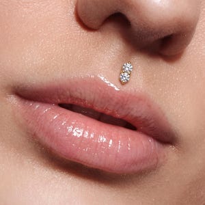 May include: A gold philtrum piercing jewelry piece featuring two vertically aligned, round, clear, faceted stones. The jewelry is positioned in the center of the skin area directly above the upper lip, showcasing a polished metal setting.