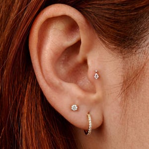 Peut inclure: Un ensemble de piercings d'oreille en or comprenant un petit clou en forme de cerf-volant avec une pierre claire au tragus, un clou rond taille brillant au lobe et une petite créole en or sertie de minuscules pierres claires.