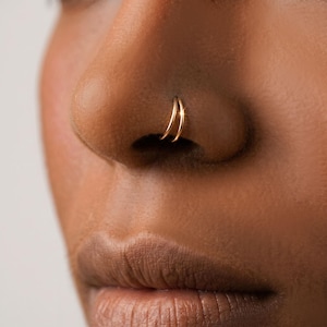Può includere: Primo piano di un naso con due sottili piercing al naso dorati. Gli anelli sono circolari e vicini, creando un effetto a doppio cerchio. L'incarnato è marrone caldo e i gioielli aggiungono un accento sottile ed elegante.