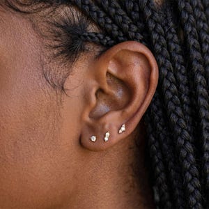 Könnte beinhalten: Drei kleine goldene Ohrstecker werden in einem Ohrläppchen getragen. Der erste Ohrstecker ist ein einzelner runder klarer Stein, der zweite ist ein vertikaler Stab mit zwei kleinen klaren Steinen und der dritte ist eine kleine Gruppe aus drei klaren Steinen.