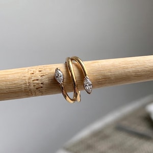 Könnte beinhalten: Ein goldfarbener Spiralring mit zwei marquiseförmigen, klaren Edelsteinen. Der Ring ist um einen Holzstab gewickelt, was sein Design hervorhebt. Die Edelsteine sind an den Enden der Spirale angebracht und verleihen dem Schmuck Glanz.