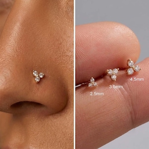 May include: A gold nose stud featuring a cluster of three small clear stones arranged in a triangular shape. The image displays three variations of the stud with labelled sizes of 2.5mm, 3.5mm, and 4.5mm, showing the jewellery on skin.