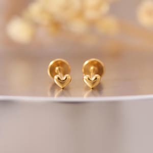 May include: A pair of gold-toned stud earrings featuring a small, smooth heart-shaped design. Each earring has a polished metallic finish and a flat circular backing, presented as a matching set of two individual heart-shaped jewelry pieces.