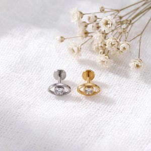 Pode incluir: Dois brincos de pino em formato de olho, um em tom prateado e outro em tom dourado. Cada brinco apresenta uma pedra preciosa redonda e transparente no centro de uma moldura oval, fixada a um suporte circular plano para fixação na orelha.
