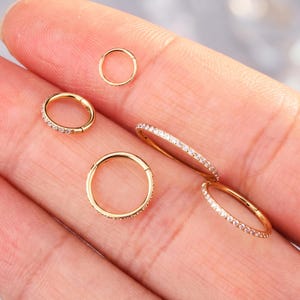 Könnte beinhalten: Eine Sammlung von fünf goldfarbenen, kreisförmigen Creolen in verschiedenen Größen. Vier der Ringe sind mit einer Reihe kleiner, klarer, funkelnder Steine am Außenrand besetzt, während ein kleinerer Ring schlicht goldfarben ist.
