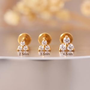 Könnte beinhalten: Drei goldene Ohrstecker, die jeweils aus drei klaren, rund geschliffenen Steinen in dreieckiger Anordnung bestehen. Die Ohrstecker sind mit den Größen 2,5 mm, 3,5 mm und 4,5 mm beschriftet, die ihre jeweiligen Durchmesser in Millimetern angeben.