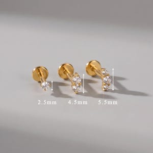 Peut inclure: Trois boucles d'oreilles à tige dorées avec des pierres rondes transparentes. La première a une pierre de 2,5 mm, la deuxième deux pierres de 4,5 mm et la troisième trois pierres de 5,5 mm. Chaque boucle possède un fermoir plat et un sertissage à griffes.