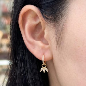 May include: A gold hoop earring featuring a row of small clear stones along the band and a dangling charm composed of three marquise-cut clear stones arranged in a fan shape, suspended from a small circular gold ring attached to the main hoop.
