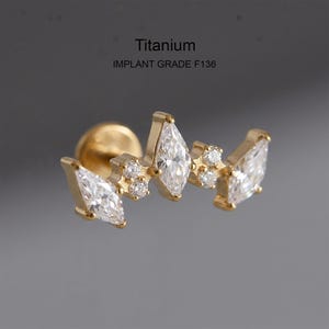 Könnte beinhalten: Ein goldfarbener Ohrstecker mit flacher Rückseite aus Titan der Implantatqualität F136, der ein geschwungenes Design mit drei klaren Steinen im Marquise-Schliff und zwei kleinen runden klaren Steinen aufweist. Das Schmuckstück hat einen sicheren Verschluss.