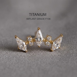 Pode incluir: Uma joia corporal em tom dourado com uma disposição em forma de coroa de três pedras claras de corte marquise e quatro pedras claras redondas. O texto TITANIUM IMPLANT GRADE F136 está impresso acima da joia, que tem acabamento metálico.