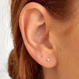 May include: A small, polished gold-tone heart-shaped stud earring is positioned in the lower earlobe. The earring features a smooth, reflective surface and a simple, symmetrical heart design that sits flush against the skin of the earlobe.