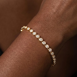 Könnte beinhalten: Ein goldenes Tennisarmband mit einer durchgehenden Reihe runder, klarer, facettierter Steine, die jeweils einzeln in einer polierten Goldfassung sitzen. Das Armband zeigt ein einheitliches Design mit einer hellen, reflektierenden Metalloberfläche.