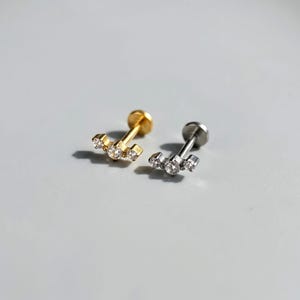 May include: Two flat back labret studs, one in gold tone and one in silver tone, each featuring a horizontal bar design set with three clear round-cut stones. The studs have a post and a circular flat back base for secure placement in piercings.