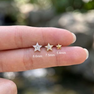 Può includere: Tre orecchini a lobo a forma di stella in tonalità oro. L'orecchino a sinistra misura 8,6 mm con una grande pietra chiara, quello centrale 7,5 mm con una pietra centrale e piccoli accenti, e quello a destra è una stella semplice da 5,4 mm.