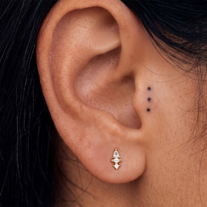 May include: A gold stud earring featuring a vertical arrangement of three clear stones, with a pear-shaped stone at the top, a marquise-shaped stone in the center, and a small round stone at the base, secured in the earlobe.