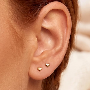 May include: Two small, polished gold-tone heart-shaped stud earrings are positioned on the earlobe. Each earring features a smooth, rounded surface and a classic heart silhouette, providing a simple and reflective metallic finish on the skin.