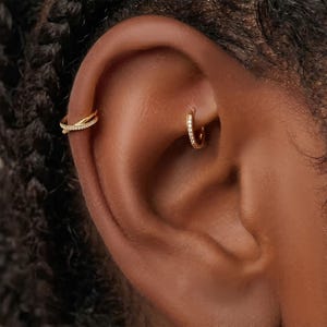 Könnte beinhalten: Nahaufnahme eines Ohrs mit zwei goldenen Ohrringen. Einer ist ein kleiner Creolen-Ohrring mit eingebetteten klaren Steinen, der andere ist ein doppeltes Ohr-Manschetten-Design mit einem Kreuzmuster, ebenfalls mit klaren Steinen. Der Schmuck ist auf dunkler Haut.