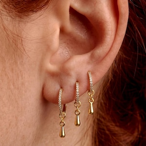 Peut inclure: Trois créoles dorées portées en rangée sur un lobe d'oreille. Chaque créole est incrustée de petites pierres claires sur le bord avant et présente une breloque dorée polie en forme de goutte d'eau suspendue à la base de l'anneau.