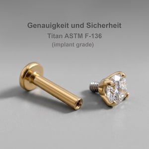 Può includere: Un gioiello da piercing tipo labret color oro con base piatta e una parte superiore separata con pietra trasparente incastonata a griffe. Il testo Genauigkeit und Sicherheit Titan ASTM F-136 (implant grade) è visibile sopra i componenti.