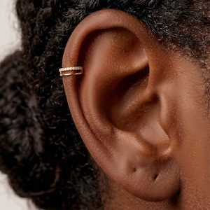 Könnte beinhalten: Ein goldfarbener Ohrklemme mit Doppelband-Design. Das obere Band ist mit kleinen, klaren Edelsteinen besetzt. Der Ohrschmuck wird am oberen Ohrknorpel getragen und verleiht dem Ohr einen Hauch von Eleganz.