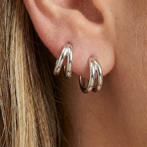 Può includere: Un paio di orecchini a cerchio argentati, ciascuno con un design a doppio cerchio. Gli orecchini sono lucidi e riflettono la luce, creando un aspetto luminoso. Sono indossati su un orecchio.