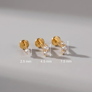 May include: Three gold-tone flat back stud earrings with clear round cubic zirconia stones. The earrings feature varying lengths of 2.5 mm, 4.5 mm, and 7.5 mm, indicated by white arrows and text labels below each piece on the display.