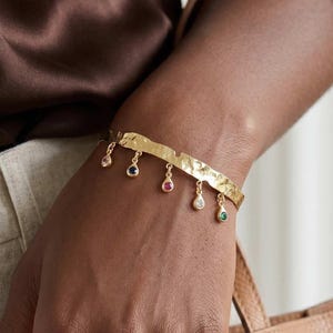 Könnte beinhalten: Ein goldfarbenes Armband mit gehämmertem Muster. Das Armband hat sechs herabhängende Anhänger in Tropfenform, jeder mit einem farbigen Edelstein: rosa, blau, rot, klar und grün. Das Armband wird am Handgelenk getragen.