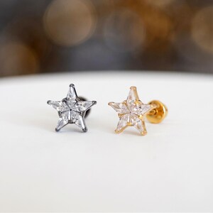 Puede incluir: Dos pendientes de botón en forma de estrella con piedras transparentes facetadas. Un pendiente tiene un engaste de metal oscuro tono plomo, mientras que el otro tiene un engaste de metal tono dorado. Ambos incluyen un cierre circular plano.