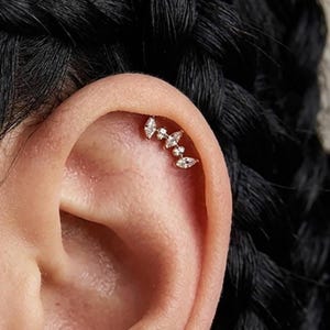 Könnte beinhalten: Ein goldfarbener Knorpel-Ohrring mit geschwungenem Design, besetzt mit drei klaren Steinen im Marquise-Schliff und zwei kleinen runden klaren Steinen in einer Reihe. Der Schmuck sitzt entlang des oberen Ohrrandes und folgt dessen natürlicher Kurve.