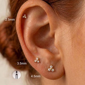 Könnte beinhalten: Drei goldfarbene Ohrstecker, die jeweils aus einer Gruppe von drei klaren Steinen in dreieckiger Anordnung bestehen. Die Ohrringe sind mit den Größen 2,5 mm, 3,5 mm und 4,5 mm beschriftet. Ein kleiner runder Einsatz zeigt den Gewindestift.