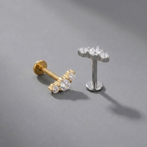 Könnte beinhalten: Zwei Labret-Piercings mit gebogenem Stab und drei in einer Reihe angeordneten, klaren, runden Steinen. Ein Piercing ist goldfarben und liegt auf der Seite, während das andere silberfarben ist und aufrecht auf seiner flachen, runden Basis steht.