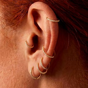 Könnte beinhalten: Nahaufnahme eines Ohrs, das mit mehreren goldenen Creolen geschmückt ist. Die Ohrringe sind mit kleinen, funkelnden Steinen verziert. Ein goldener Ohrring ist ebenfalls oben am Ohr zu sehen. Der Hautton ist hell mit rotbraunem Haar.