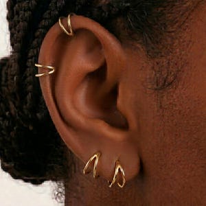 Puede incluir: Primer plano de una oreja adornada con múltiples pendientes de aro y puños dorados. La joyería incluye aros pequeños y delicados y puños de doble banda, creando un look elegante y en capas. El tono de piel es marrón cálido.