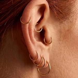 May include: Close-up of an ear adorned with multiple gold hoop earrings of varying sizes. The earrings are simple, circular designs, creating a layered effect. The skin tone is light, and the focus is on the jewelry.