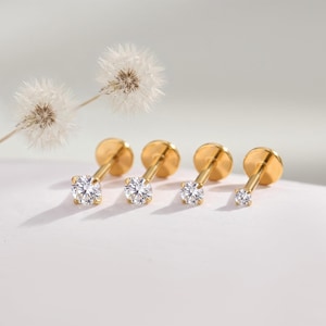Può includere: Quattro orecchini a lobo color oro con retro piatto, ciascuno caratterizzato da una singola pietra preziosa rotonda trasparente incastonata a griffe, disposti in fila accanto a due soffici fiori di tarassaco bianchi.