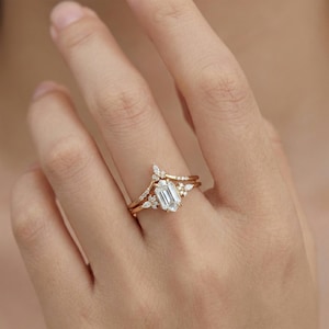 Puede incluir: Un juego de anillos de compromiso de oro con una piedra central de talla esmeralda y una banda delicada. El conjunto incluye una banda curva con pequeñas piedras de acento, creando un aspecto único y elegante. El anillo se muestra en un dedo.