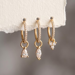 Op de afbeelding: Drie gouden oorringen met pavé-gezette steentjes langs de ring. Elke ring heeft een hangende bedel: de eerste met een peervormige steen, de tweede met een marquise-geslepen steen en de derde met een cluster van drie kleine ronde steentjes.